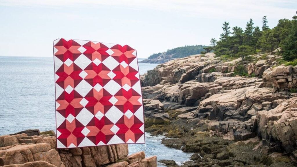 Quilt photo taken by guest teacher Kitty Wilkin on a rocky shoreline. Quilt fabrics are red, coral and maroon on a white background.