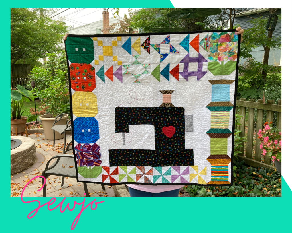 Quilt design with thread spools and button blocks surrounding a pieced sewing machine. Made by Karen Sykes, Make Modern Triangles member spotlight.